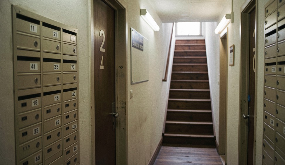 Narrow guesthouse corridor with numbered doors, a wall of numbered mailboxes and a staircase sign lit by a fluorescent tube